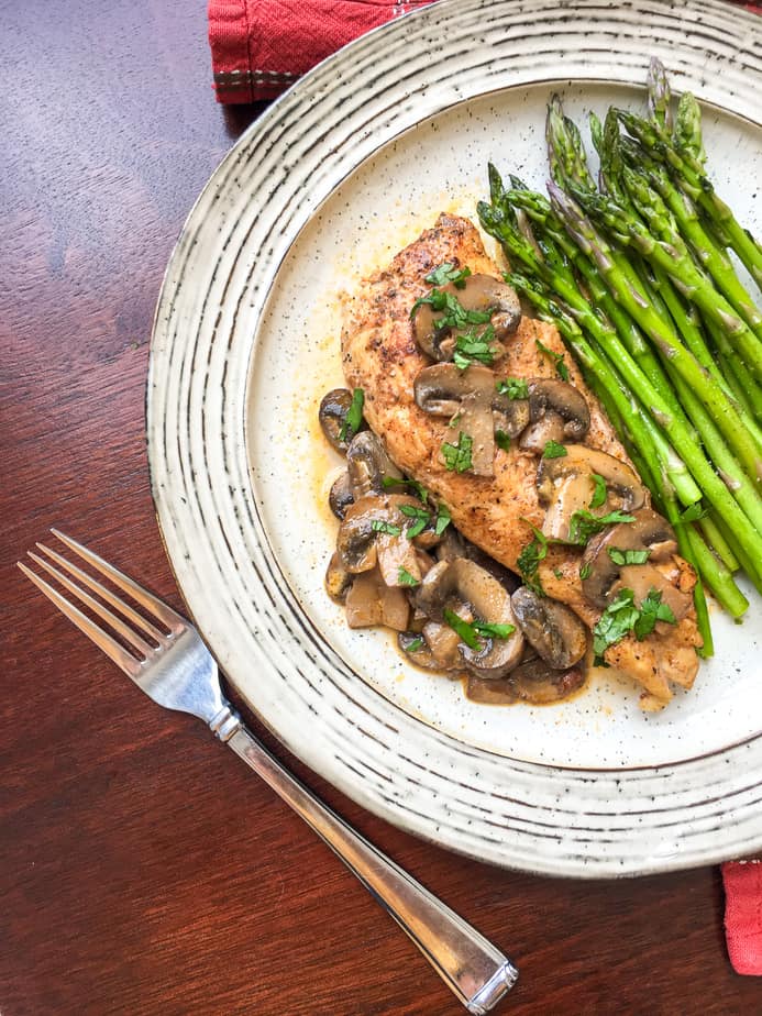 off-white plate with chicken covered in mushrooms and a brown sauce with a side of asparagus