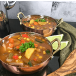 Vegetable soup in 2 copper bowls on a black table with 2 lime wedges between the two bowls