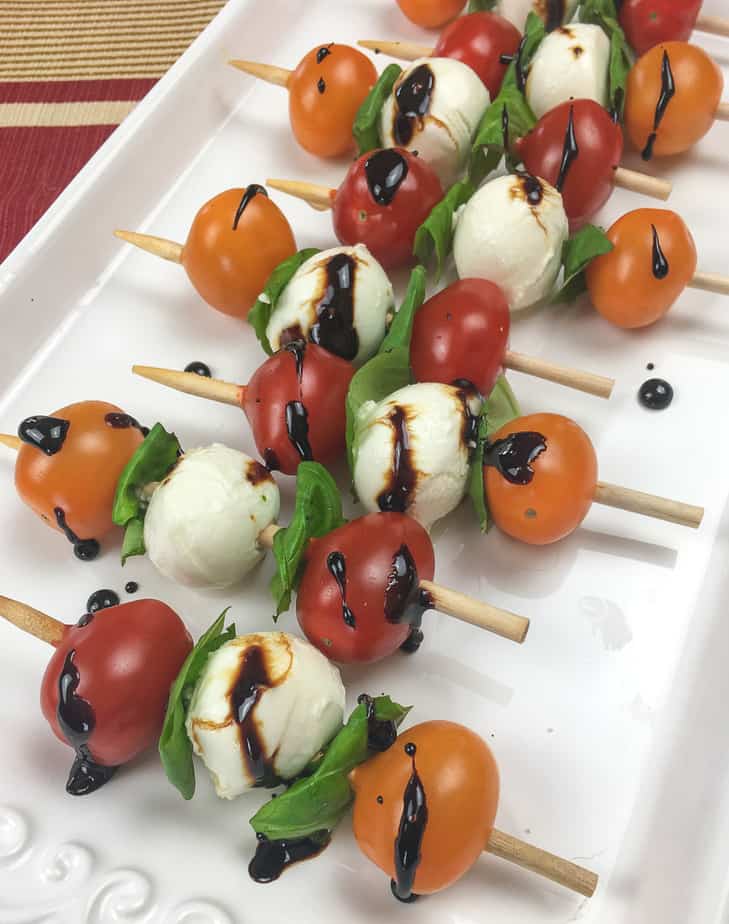 Six skewers with orange and red tomatoes, basil, small balls of mozzarella cheese and a drizzle of balsamic glaze sit on a a white platter. 