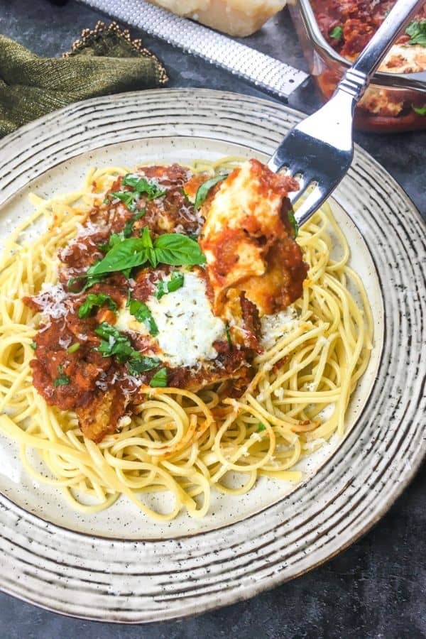 On a white plate, there's a bed of pasta. On the pasta site a piece of chicken parmesan with cheese and basil on top. There is a fork hovering above the plate with a piece of chicken on it. 