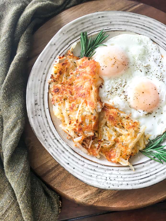 There's a wooden cutting board, with a grey cloth next to it. On the cutting board is a white plate with speckles on it. On t he plate are hash browns, sunny-side up eggs and a sprig of rosemary.
