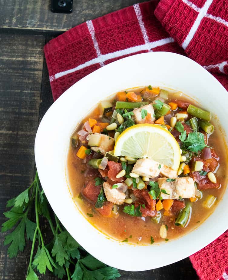 On a wooden table sits a red napkin with a waffle texture and white lines on it. On that sits a white bowl filled with fish stew, topped with a lemon slice. Next to the bowl are some fresh herbs. 