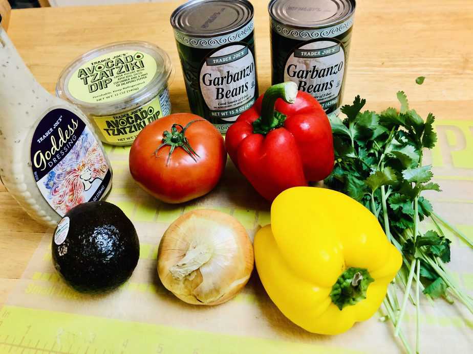 Trader Joe's Green Goddess dressing, Trader Joe's avocado tzatziki dip, 2 cans of garbanzo beans, an avocado, a tomato, a red pepper, a yellow pepper, an onion, cilantro, all on a green cutting board. 