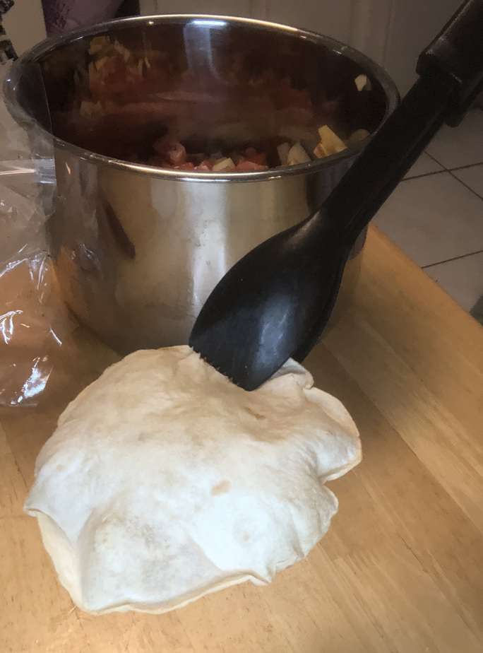 A silver pot with some tomatoes in it. In front of that is a tortilla that's puffed up, held by black tongs. 