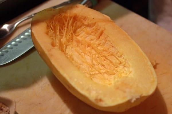 Half of a spaghetti squash on a wooden cutting board, with a carving knife  and a spoon in the background.