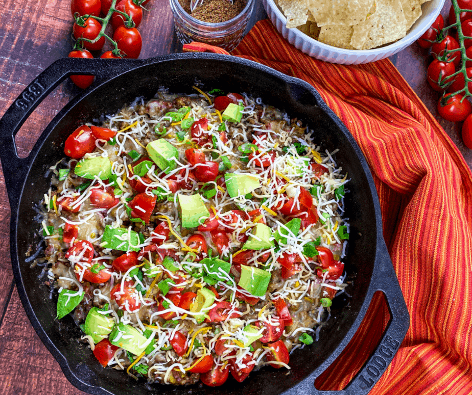 A cast-iron skillet with beef on the bottom and covered with cherry tomatoes, chunks of avocado, and a sprinkling of cheddar cheese. On the top left there are cherry tomatoes on the vine. Next to that is a small glass bowl with a spoon in it and some spices. To the right of that is a white bowl with corn chips in it. Everything is on a wooden table. There's a red napkin with orange and yellow thin stripes on it to the right of the skillet.