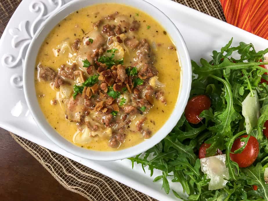 A white bowl sits on a white plate. Lower left there's a salad with greens and tomato. In the bowl is an orange soup with bacon and parsley on top. There's a spoon in. the foreground, held over the bowl, with soup in it
