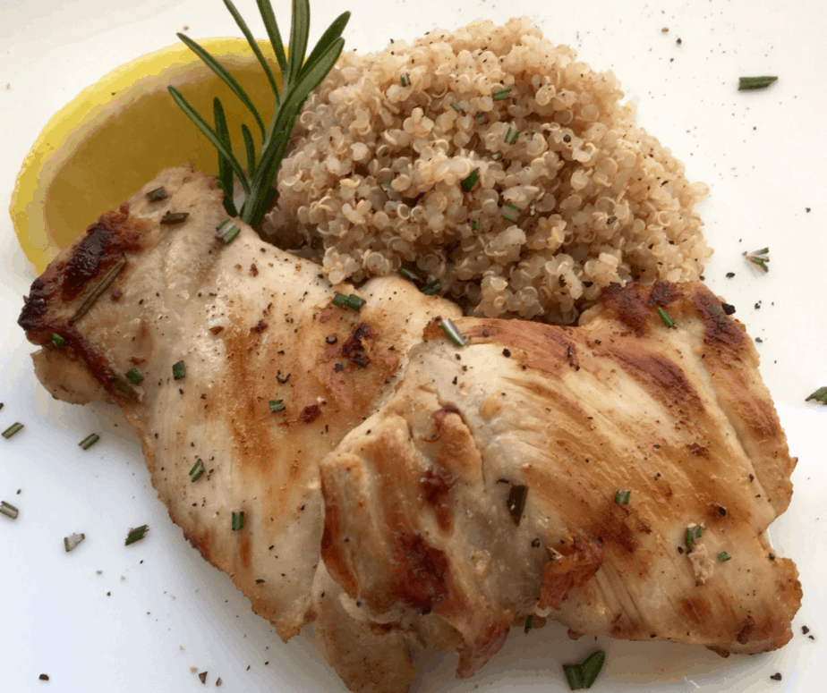 A nicely browned chicken thigh sits on a white plate. To the right is a pile of rice, on top of the rice and chicken is a sprig of rosemary and a lemon wedge. 