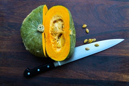 An acorn squash is cut in half, on a wooden table. The knife is under it and there are seeds on the table. 