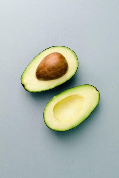 An avocado cut in half, sitting on a light grey table. There is a pit on the half on the top.