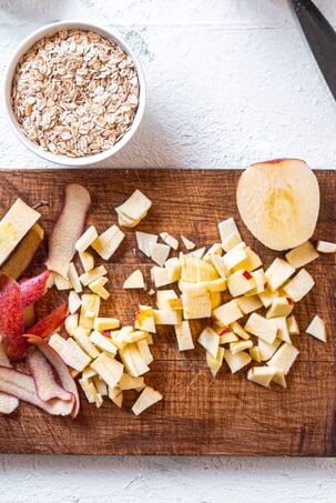 A wooden cutting board with diced apples, apple peels, and half an apple. To the top left is a small white bowl with uncooked oats