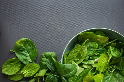 On a grey table, there's a metal bowl filled with spinach leaves, with leaves on the table to the left of the bowl.