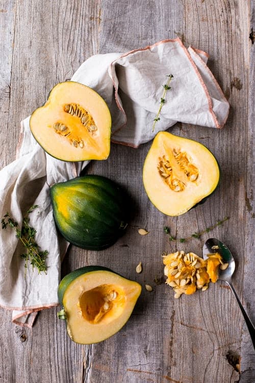 On a wooden table, there is a white napkin with red stitching on the border. Half an acorn squash is on the napkin, another half beside that. There's a whole acorn under the halves and a half under the whole. That half hals seeds scooped out of it. The seeds are next to the half, on the table, with a spoon next to them. 