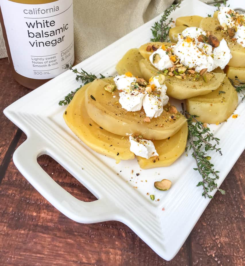 Golden Beets arranged on a white platter garnished with thyme and pistachios a bottle of white balsamic vinegar in the background