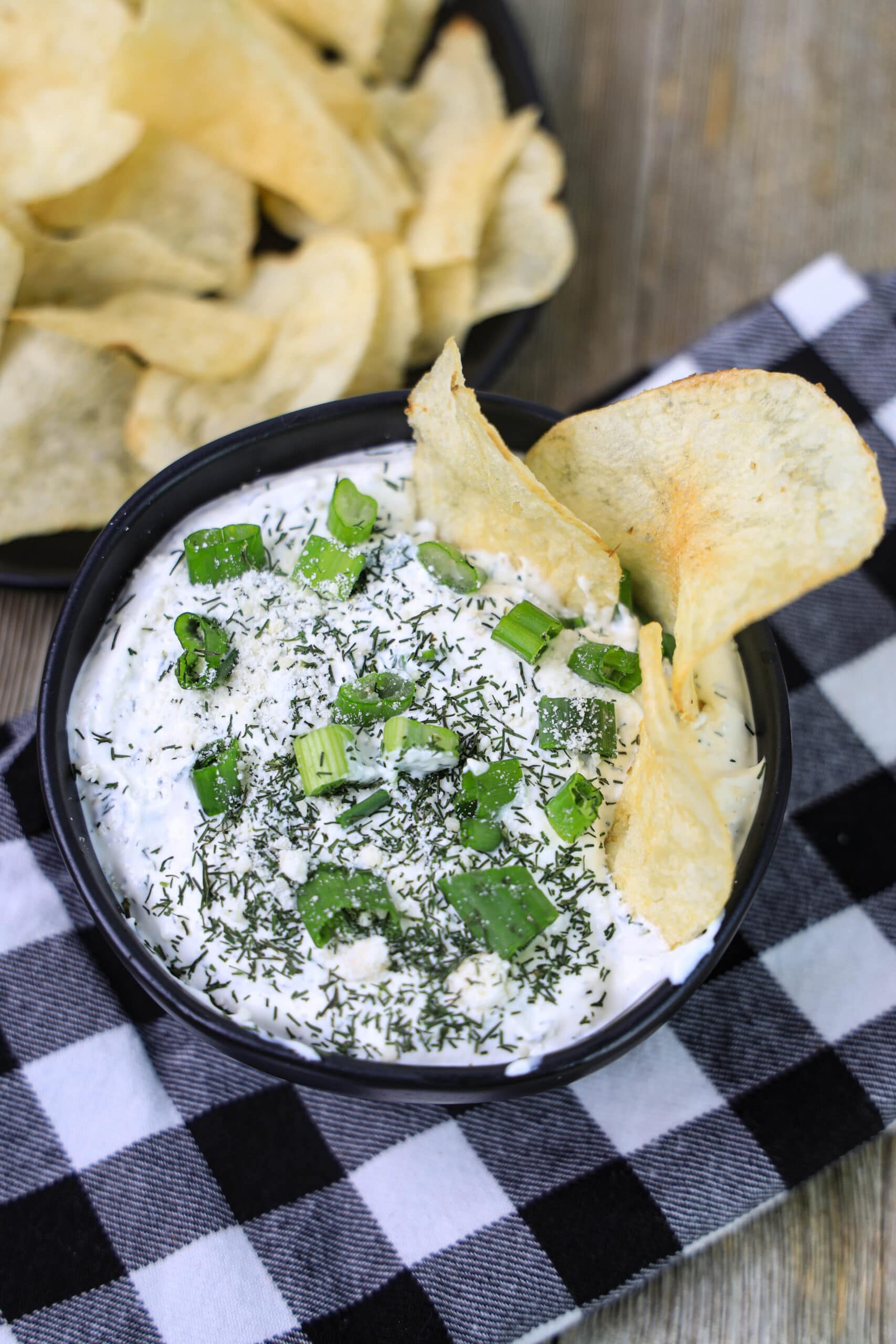 dill dip in a black bowl with potato chips and a side bowl of potato chips on a wood table.