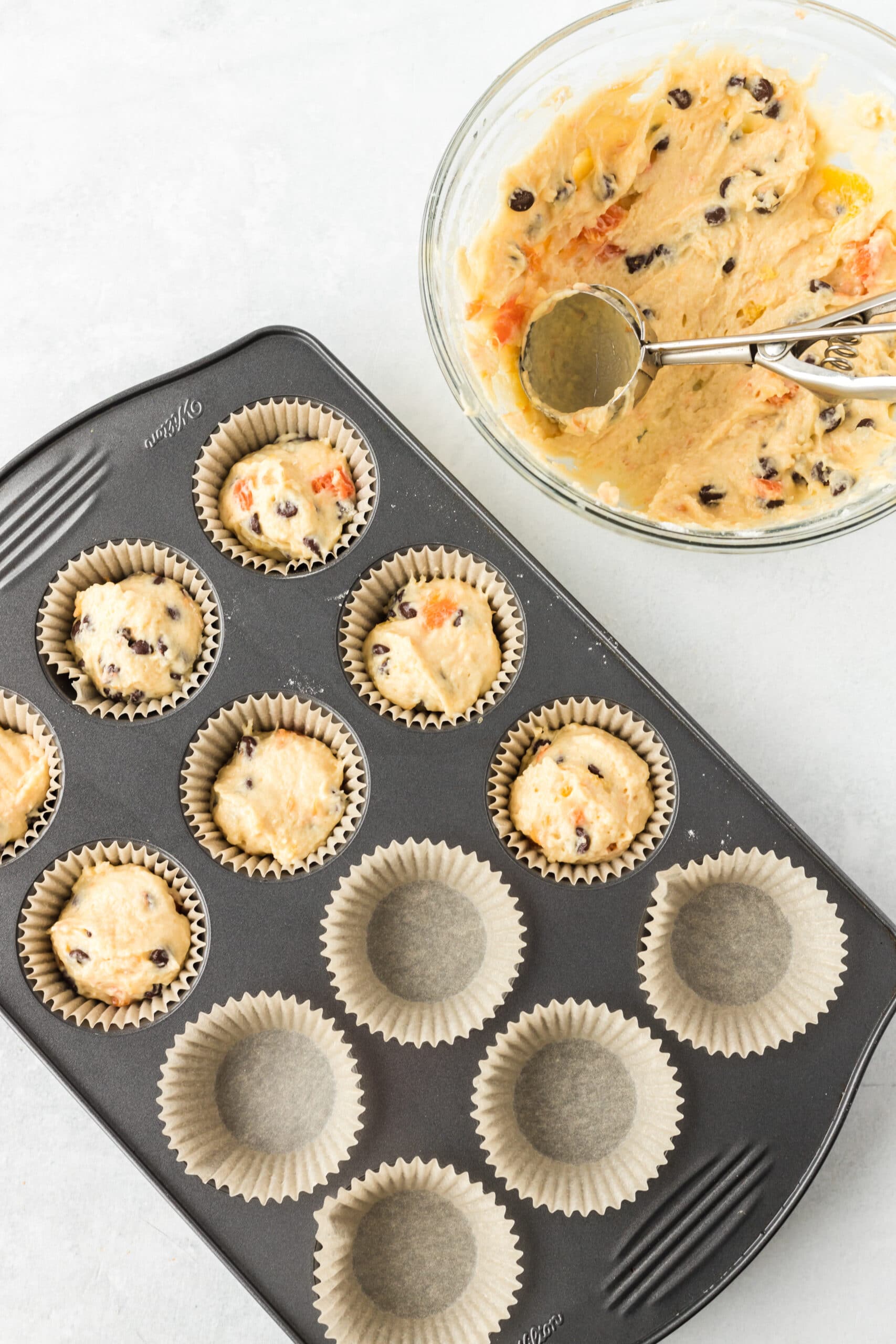 A clear bowl with orange muffin batter in it. Next to that is a muffin pan partially filled with batter on a white table.