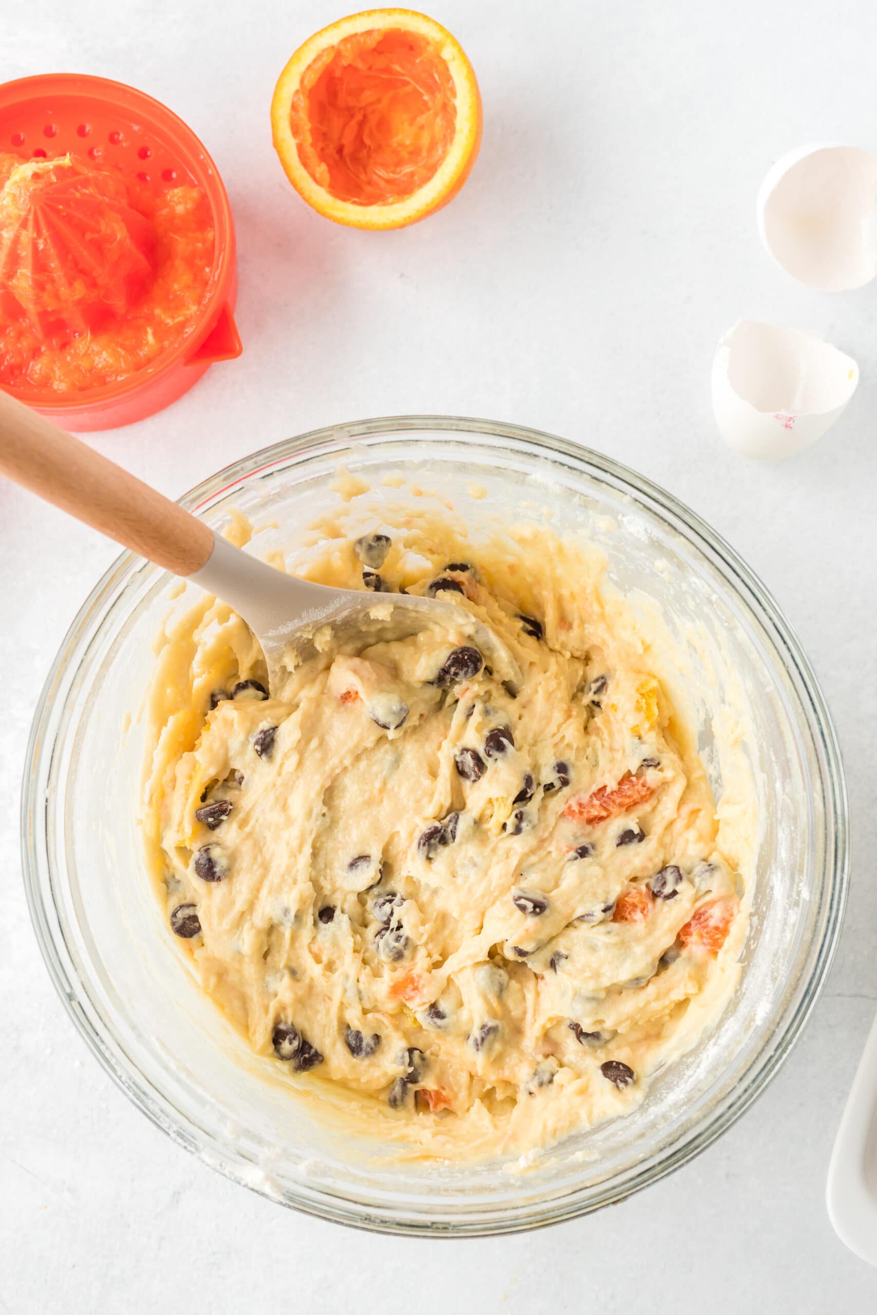 An orange that has been juiced next to a hand juicer and a clear bowl with chocolate chips and batter in it, being mixed with a wooden spoon