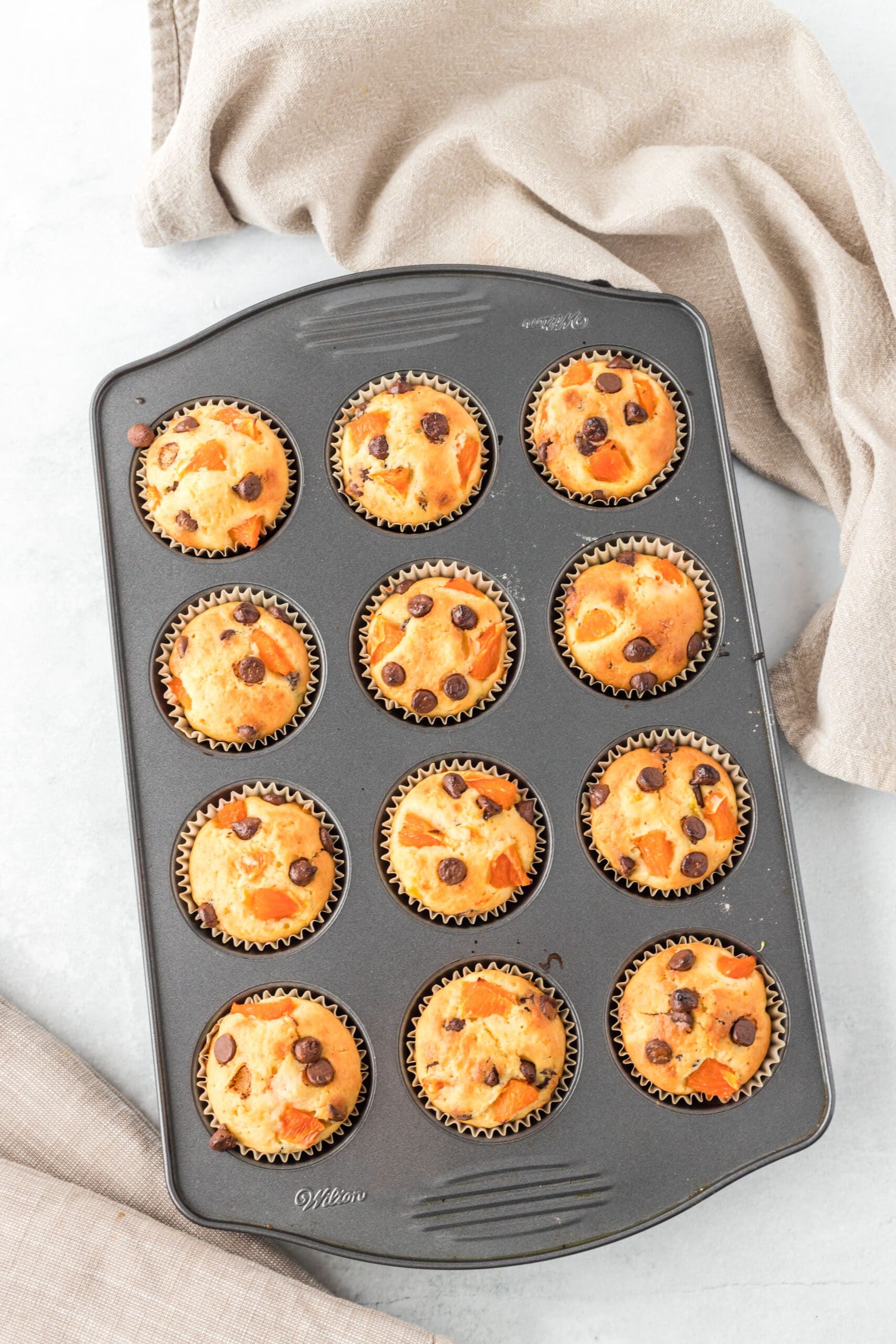 orange breakfast muffins in a muffin pan on a white table with a white towel near the top of the picture.