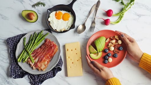 Ketogenic diet meal preparation female hands serving red plate with avocado, blueberry, Brazil nut, macadamia, pecan, walnut on marble table with fried eggs, bacon, roasted asparagus and cheese.