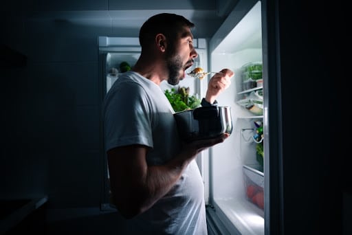 Hungry man eating food at night from open fridge