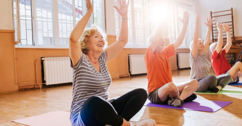 A group of people are gathered in an indoor space, practicing yoga. All four figures have their feet firmly planted on the ground while they practice their poses together